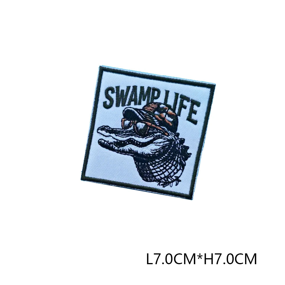 Black embroidered patch with white text SWAMP LIFE featuring a stylized fish head