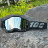 A pair of black motocross goggles with a reflective lens and a gray strap resting on a large rock.