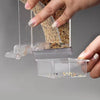 A hand is holding a transparent anti-spill parrot feeder containing seeds and grains.