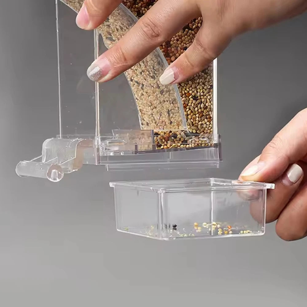 A hand is holding a transparent anti-spill parrot feeder containing seeds and grains.