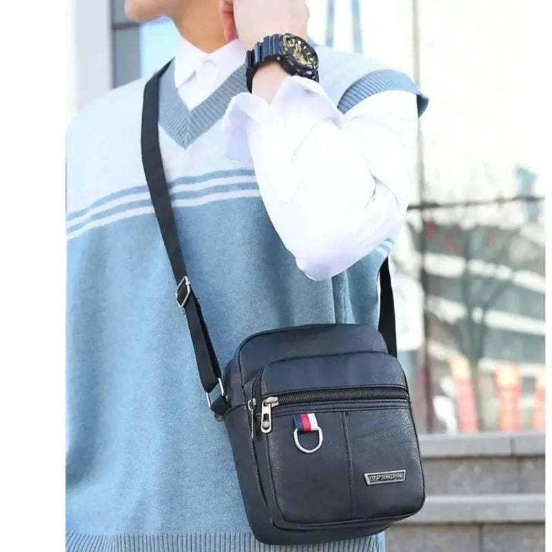 Un homme portant un pull bleu clair et une chemise blanche tient un sac bandoulière noir en selle.