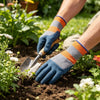 A hand wearing orange and gray gardening gloves is using a small metal trowel to dig in dark soil among plants.