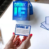 A hand holds a card from the Drink IF adult drinking game deck, featuring text about congratulating someone for being pregnan
