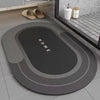 A round gray bath mat with a dark oval design and non-slip rubber base is displayed on a tiled floor.