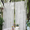 A white floral lace sheer curtain panel hanging outdoors between tree trunks.