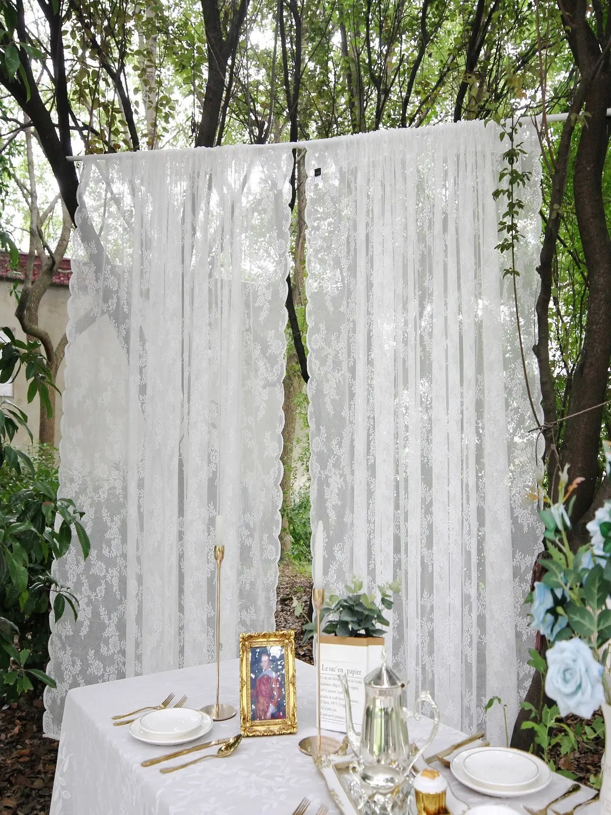 A white floral lace sheer curtain panel hanging outdoors between tree trunks.