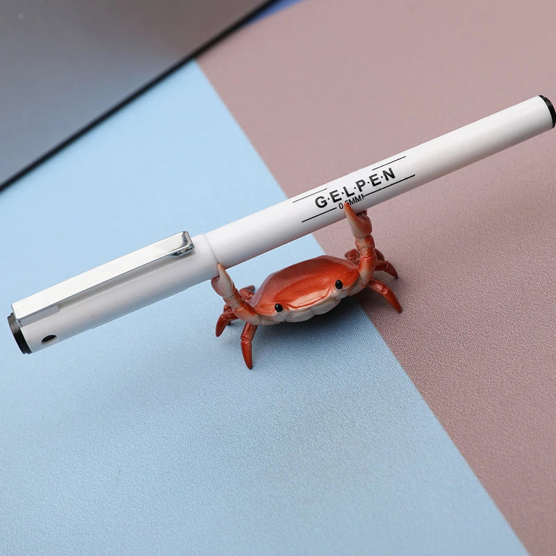 A white Gelpen pen is resting on a pink and blue surface next to a small, realistic-looking crab.