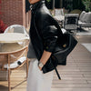 A woman wearing a black leather jacket and white pants holds a black leather square crossbody bag with a gold lock.