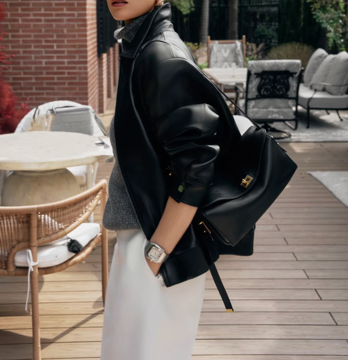 A woman wearing a black leather jacket and white pants holds a black leather square crossbody bag with a gold lock.