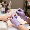 Hands wearing light purple disposable nitrile gloves applying a dark cosmetic product to a finger.