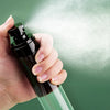 A hand is holding a dark glass spray bottle, releasing a fine mist of white powder against a green background.