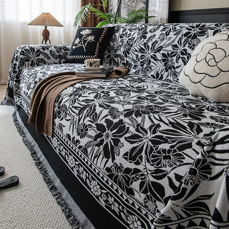 A black and white floral jacquard sofa blanket with tassels draped over a dark upholstered sofa.