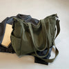 A olive green corduroy tote bag with a leather strap is shown next to a black jacket and a notebook.