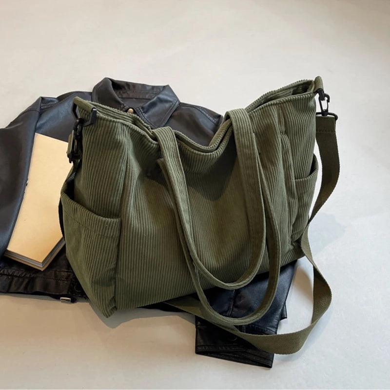 A olive green corduroy tote bag with a leather strap is shown next to a black jacket and a notebook.