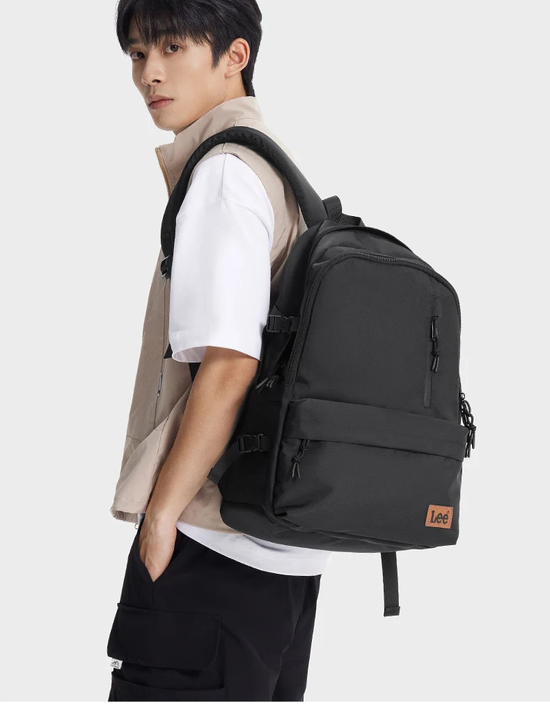 A young man wearing a beige shirt and black pants is holding an olive green or black ultralight water-resistant laptop backpa
