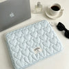 A light blue quilted laptop sleeve resting on a white surface next to a silver laptop and perfume bottle.