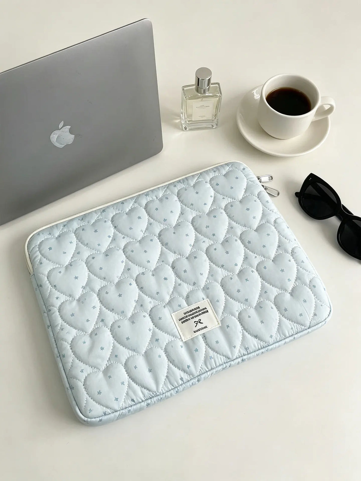 A light blue quilted laptop sleeve resting on a white surface next to a silver laptop and perfume bottle.