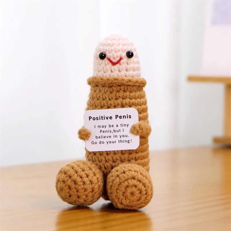 A handmade crochet doll resembling a positive energy figure with a pink head and brown body sitting on a wooden surface.