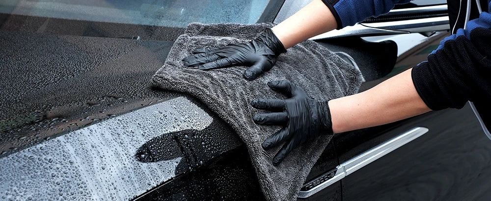 Hände in schwarzen Handschuhen reinigen ein Auto-Fenster mit einem grauen Mikrofasertuch.