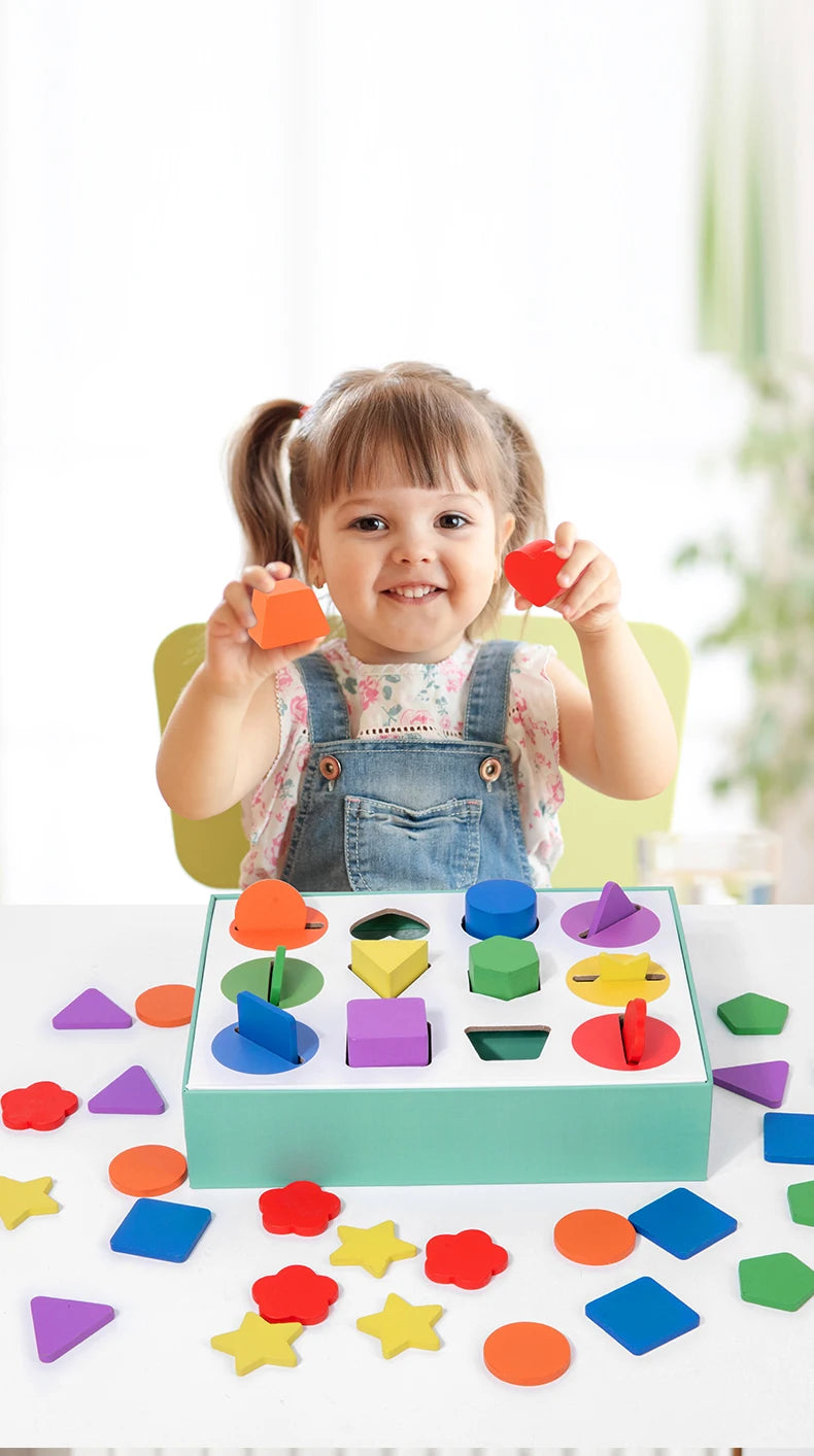 A smiling toddler holds red geometric shapes above a green wooden sorting box filled with various colored wooden shapes.
