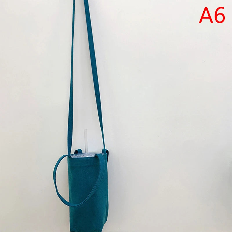 A teal canvas crossbody bag with a strap hanging against a white wall.