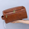 A brown leather toiletry bag with brass zippers is held by a hand against a light gray background.