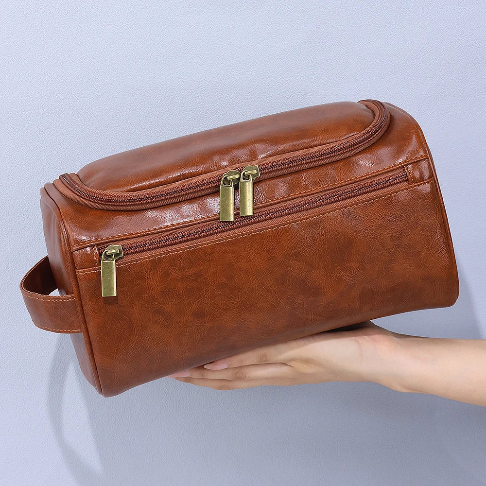 A brown leather toiletry bag with brass zippers is held by a hand against a light gray background.