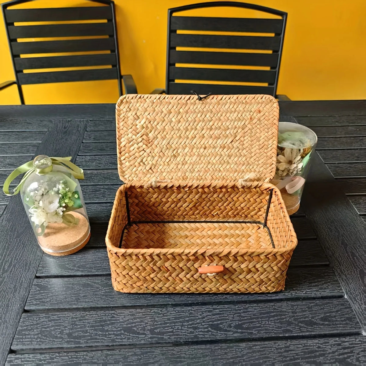 A woven rattan storage basket with a lid is displayed on a dark wooden table against a yellow wall.
