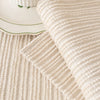 A close-up of a beige ribbed knit cushion cover resting on a light rug next to a white ceramic dish.