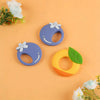 A set of silicone baby rattles and teethers in purple, blue, and yellow with flower accents on a light orange background.