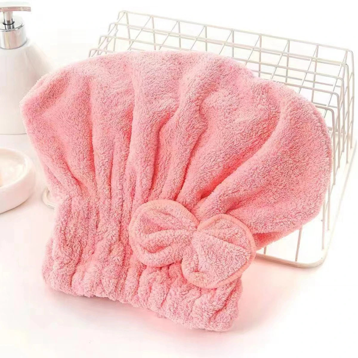 A pink microfiber hair drying towel with a bow-knot head wrap is displayed in a wire basket.
