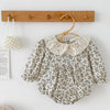 A light floral cotton romper with a delicate lace collar hanging on a wooden hanger against a white wall.