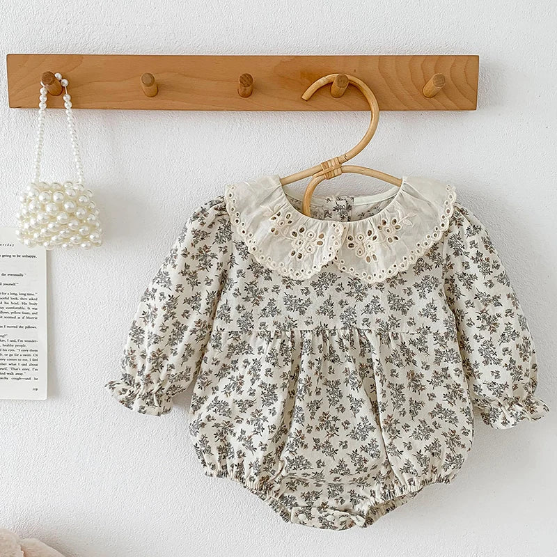 A light floral cotton romper with a delicate lace collar hanging on a wooden hanger against a white wall.
