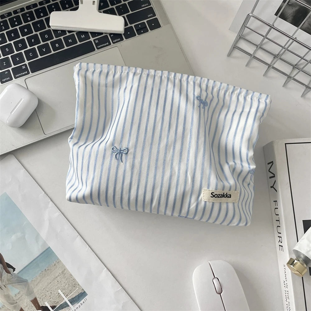 A light blue and white striped makeup bag with a floral embroidery detail resting on a desk next to a laptop and mouse.