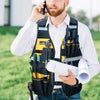 A man wearing a white shirt and dark vest with multiple tool pockets is talking on a phone outdoors.
