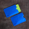 A blue RFID blocking card sleeve is shown resting on a dark brown surface next to a green card.