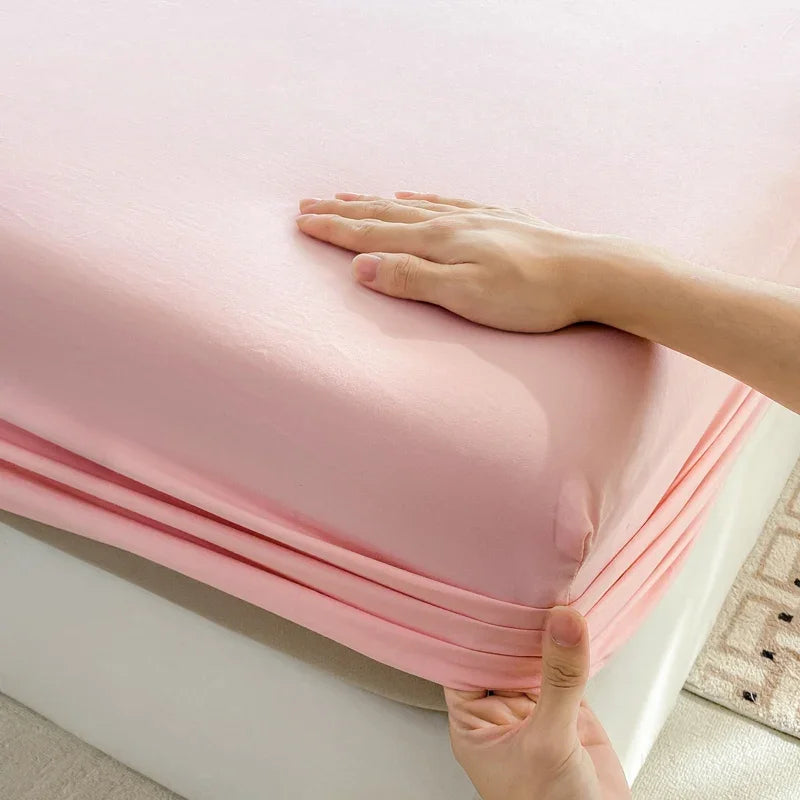 A hand is folding a pink fitted sheet with an elastic band to show its deep pocket design.