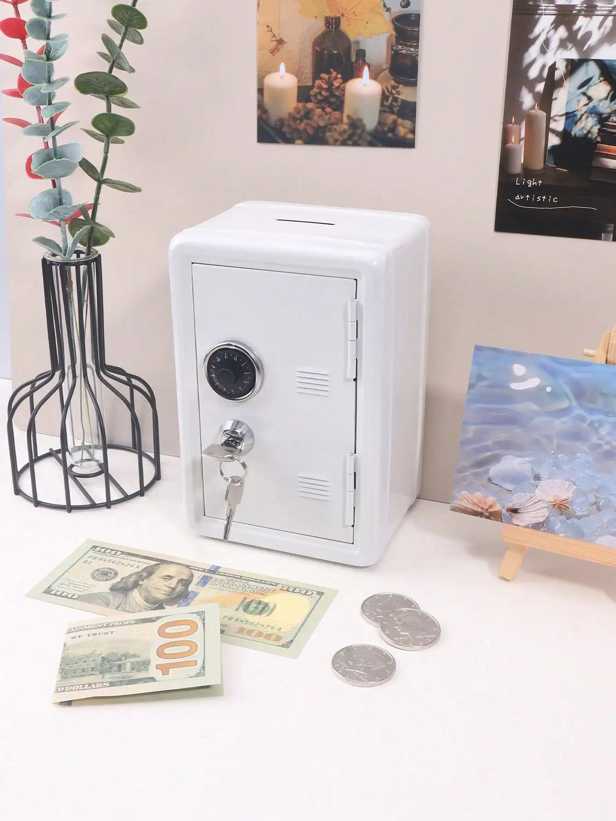 A white compact desktop safe with a combination lock sits on a white surface next to some cash and decorative items.