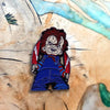 An enamel pin featuring Chucky the doll with red hair and a stitched smile against a light background.
