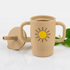 A beige silicone training cup with a straw and handles is shown on a white surface.