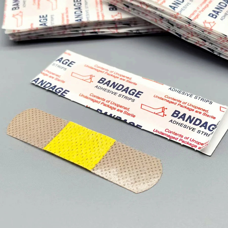 A single beige adhesive bandage strip is shown on a gray surface, with a stack of similar bandages in the background.