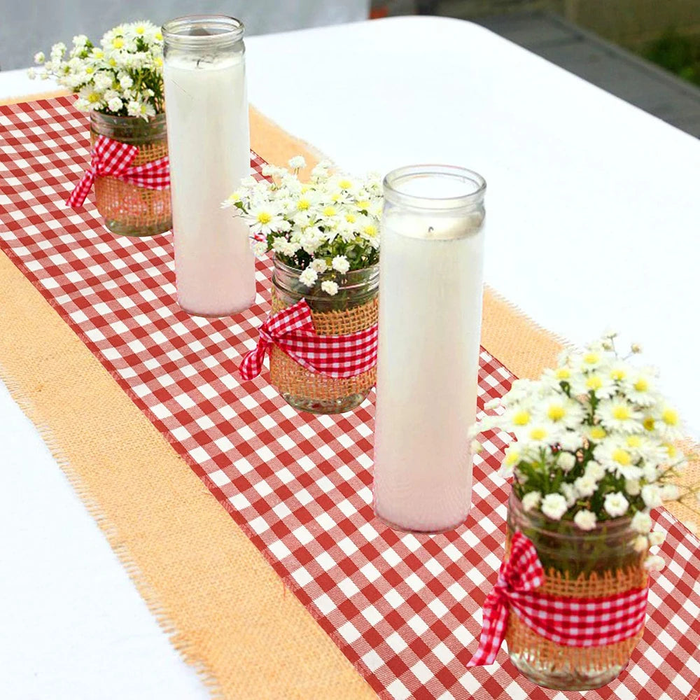 A display of white pillar candles in glass jars set on a red and white gingham table runner.
