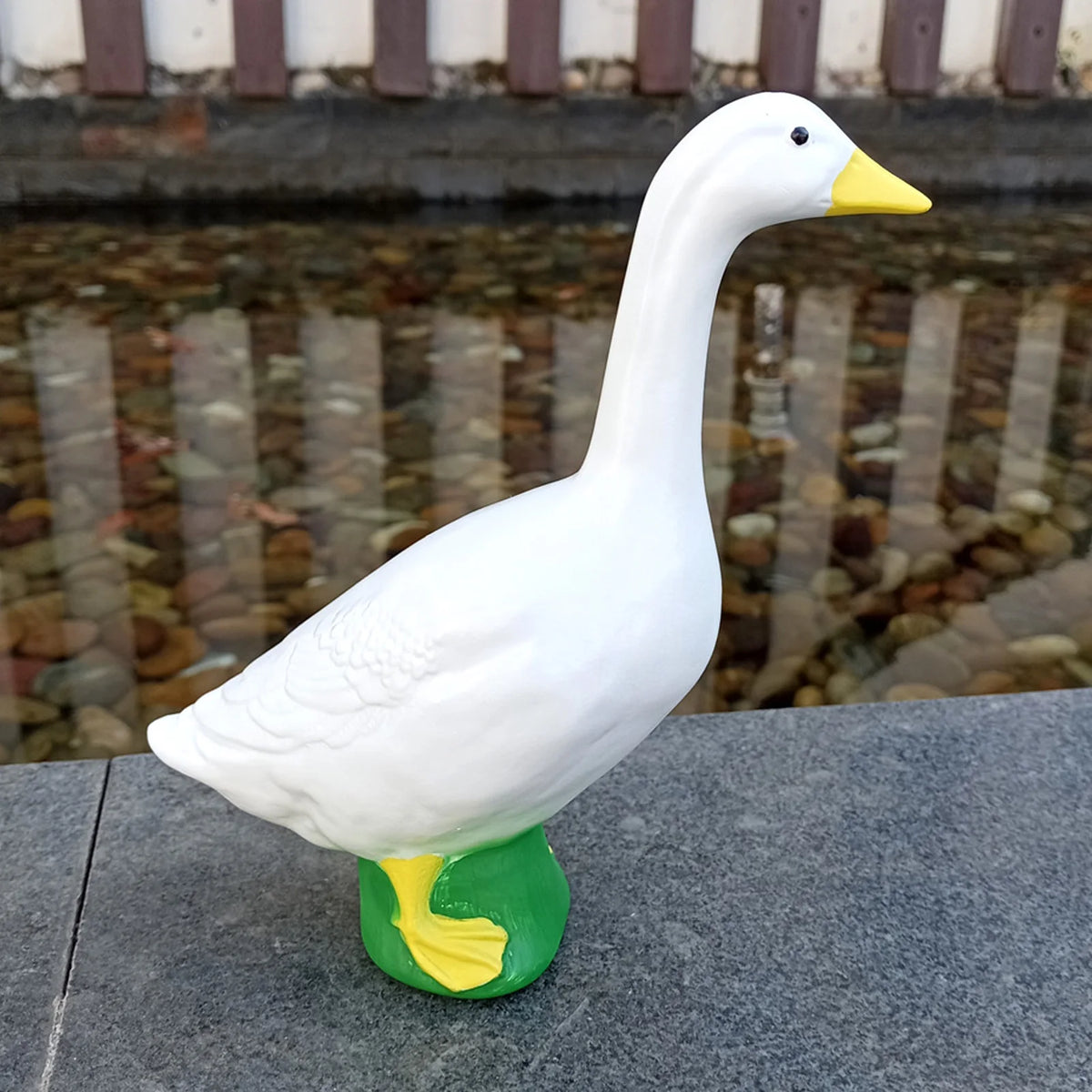 A white resin goose garden statue with a yellow beak and green base sitting on a dark surface.