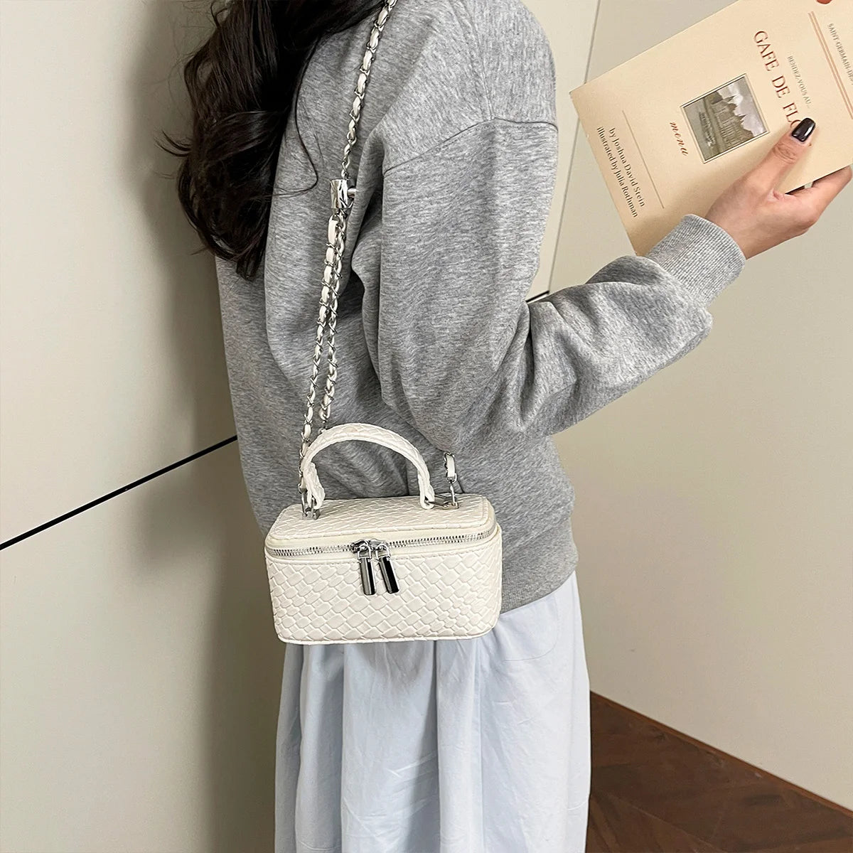 A person holds a cream woven PU leather crossbody bag and a book, against a light wall.