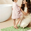 A young girl stands on a green textured bathroom rug while an adult woman holds a pink towel near a white bathtub.