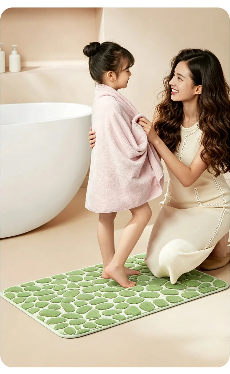 A young girl stands on a green textured bathroom rug while an adult woman holds a pink towel near a white bathtub.