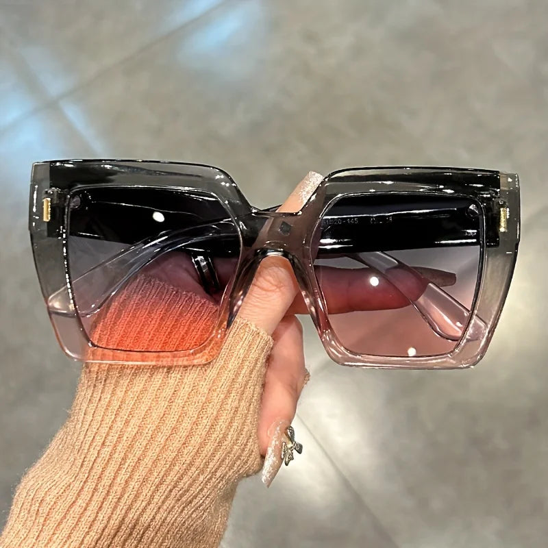 A hand holds a pair of gradient square sunglasses with pink and orange lenses against a blurred gray background.