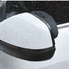 A close-up of a black carbon fiber car side mirror rain and sun visor installed on a vehicle.