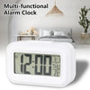 A white multi-functional alarm clock with a digital display is shown on a bedside table.