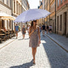 A woman in a patterned dress holds a large white umbrella while walking down a cobblestone street.